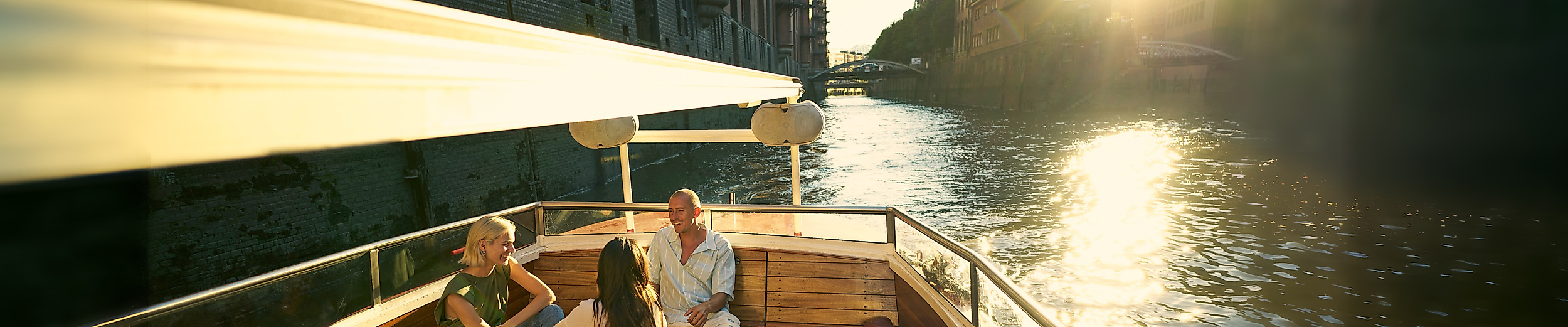 Drei Personen auf einer Barkasse bei Sonnenuntergang mit der Speicherstadt im Hintergrund