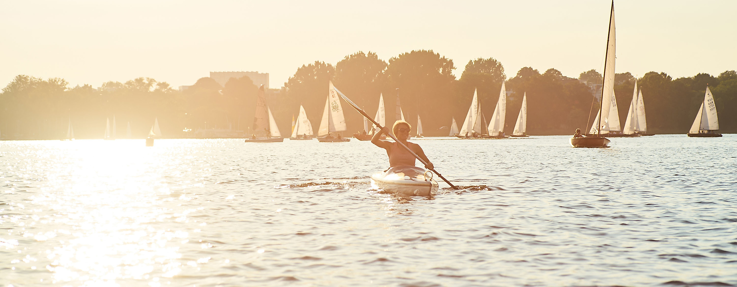 Person paddelt im Kanu auf der Alster bei Sonnenuntergang, umgeben von zahlreichen Segelbooten