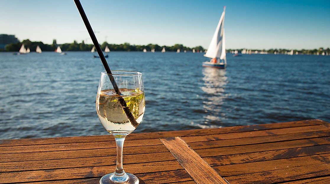 Cocktailglas mit Strohhalm auf Holzsteg an der Außenalster, im Hintergrund segeln Boote bei Sonnenschein