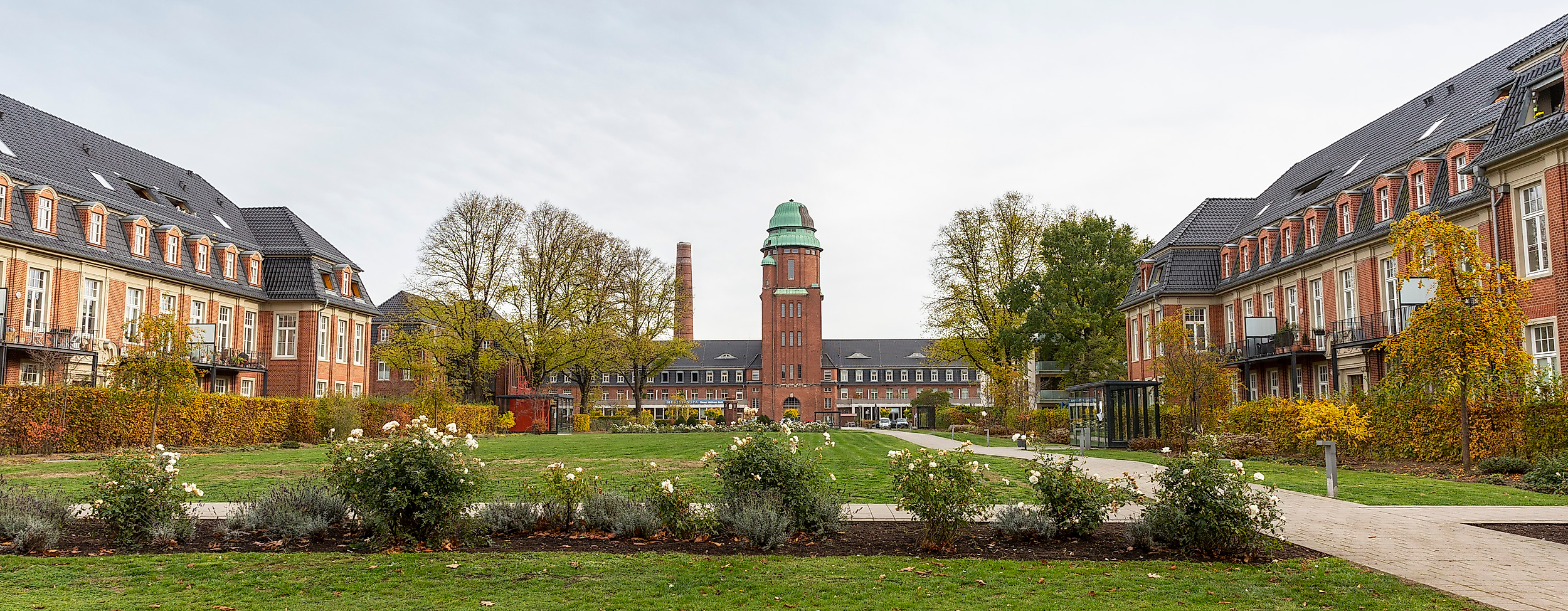 Symmetrischer Innenhof mit Rasen und Blumen in Hamburg-Dulsberg, im Hintergrund Backsteinbauten mit Uhrenturm.