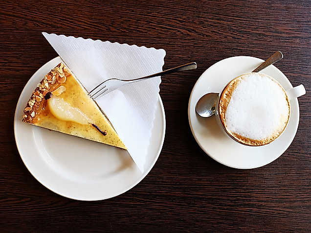 Stück Birnentarte mit Puderzucker und Cappuccino auf Holztisch, serviert in der Biokonditorei Eichel