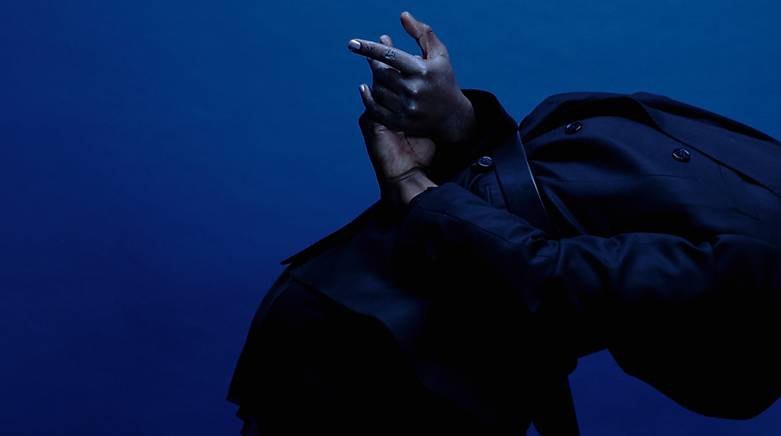 A performer is seen from the side, leaning forward, a black hoodie pulled over their head. Their hands are behind their back and stretched out.