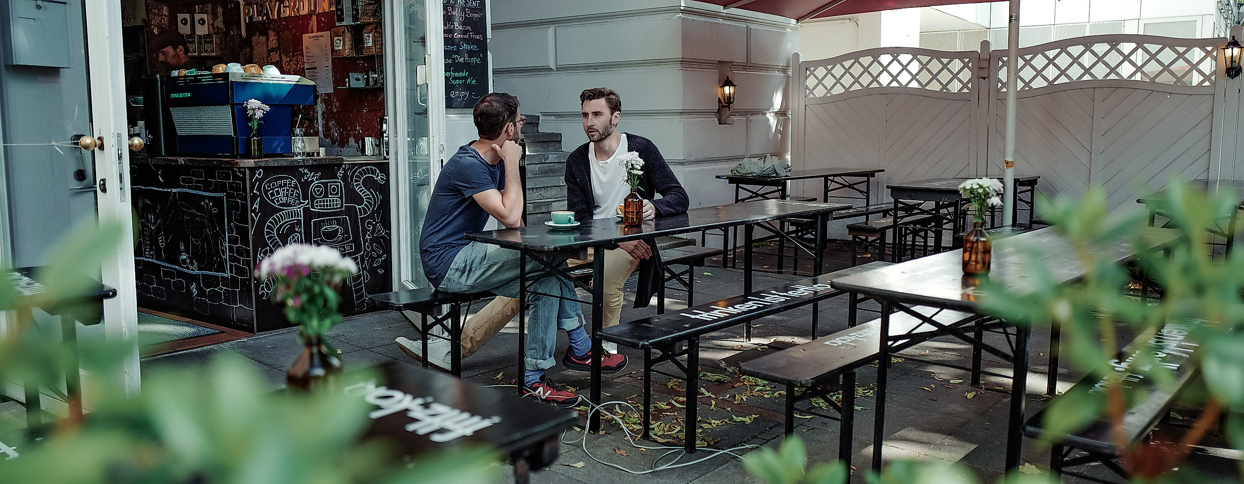 Zwei Personen sitzen im Außenbereich von Playground Coffee, umgeben von Pflanzen und roten Sonnenschirmen