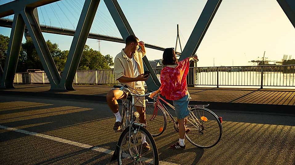 Zwei Personen mit Fahrrädern stehen auf einer Brücke in Hamburg, eingerahmt von der Stahlkonstruktion des Bauwerks. Die Szene ist in warmes Licht getaucht – es ist Sonnenuntergang. Die beiden unterhalten sich entspannt, während im Hintergrund Wasser, Hafenanlagen und Teile der Stadt sichtbar sind. Das Bild vermittelt ein Gefühl von Freiheit und urbaner Mobilität. Es symbolisiert die Vorteile der Hamburg CARD, insbesondere für Fahrradausflüge und die individuelle Erkundung der Stadt.