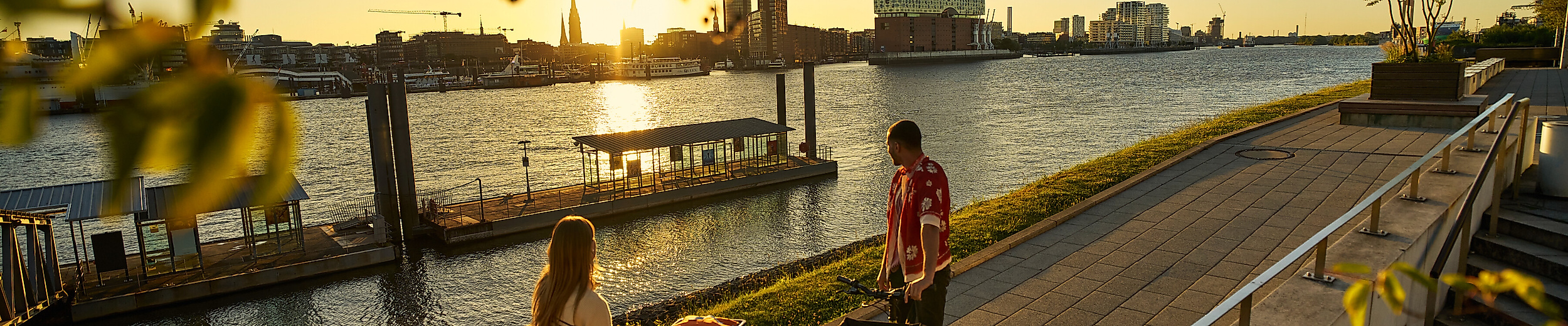 Zwei junge Erwachsene mit Fahrrädern an der Elbe mit Blick auf die Elphi.