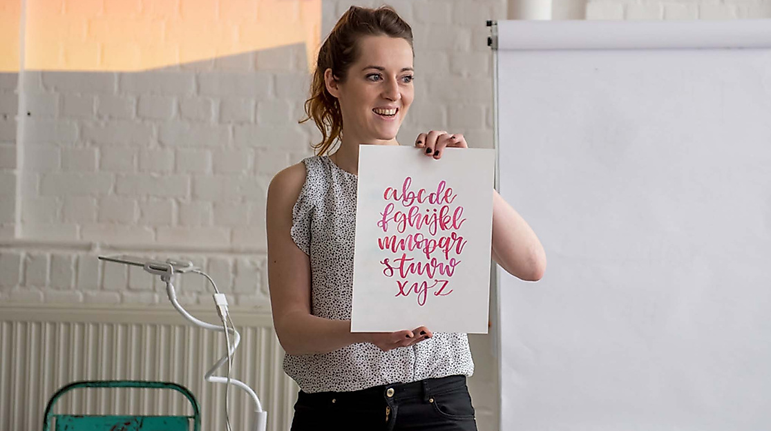 Workshop-Leiterin präsentiert gestaltetes Lettering mit pinken Buchstaben vor Flipchart in kreativem Raum