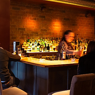 Gäste an der beleuchteten Bar der Boilerman Bar, Barkeeper mixt Cocktails vor Backsteinwand