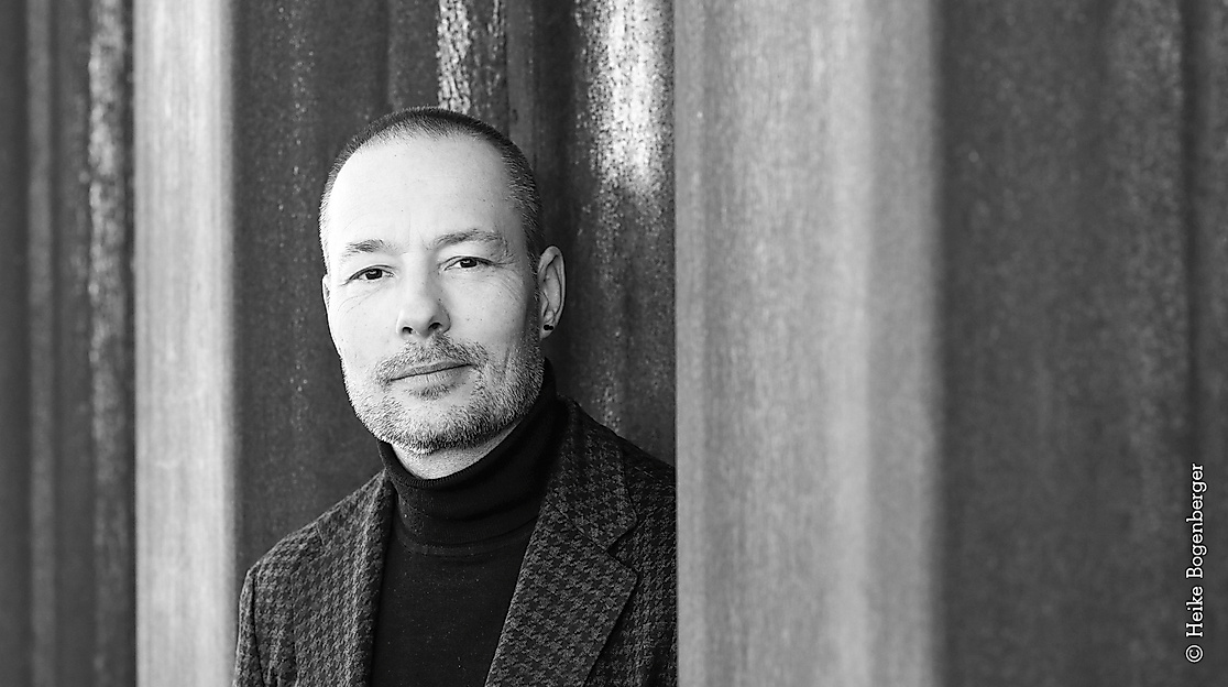 Black-and-white photograph of a male-read person with short-cropped hair, a black earring, a beard, and dressed in a plaid jacket.