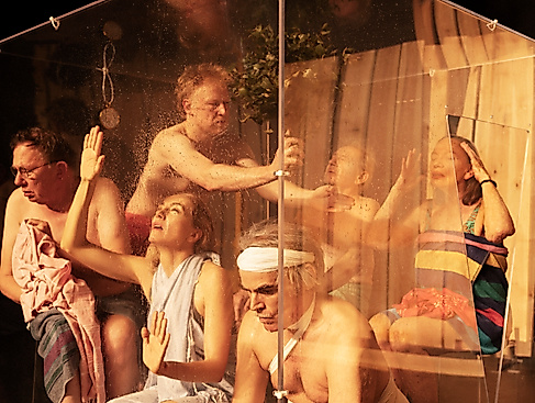 Sechs Performende sind in einer Sauna-Situation hinter einer Glaswand. Sie alle sind in unterschiedlichen Positionen und leicht bekleidet.