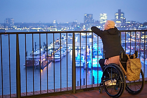 Rollstuhlnutzerin blickt abends von barrierefreier Plattform auf die beleuchtete Elbphilharmonie und den Hafen.
