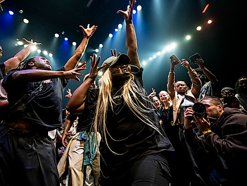 A dynamic scene in which a crowd of people stand around a dancer, cheering and cheering her on with gestures. The dancer raises her arms upwards, spreading her fingers. At the bottom right, someone photographs the dancer from close up.