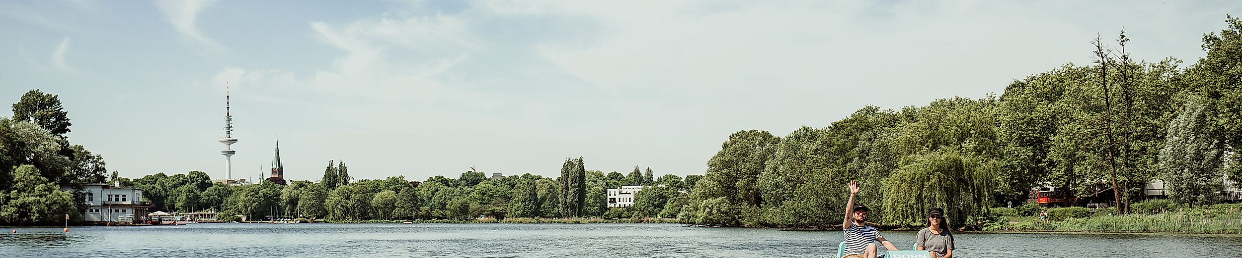 Paar fährt bei Sonnenschein mit einem Tretboot über die Alster, umgeben von Bäumen und Wasser