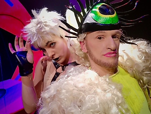 Two performers pose in costumes. One person wears a green costume with a light-colored curly wig and a headband with a large eye. The person behind them wears a harness outfit, but is only half visible.