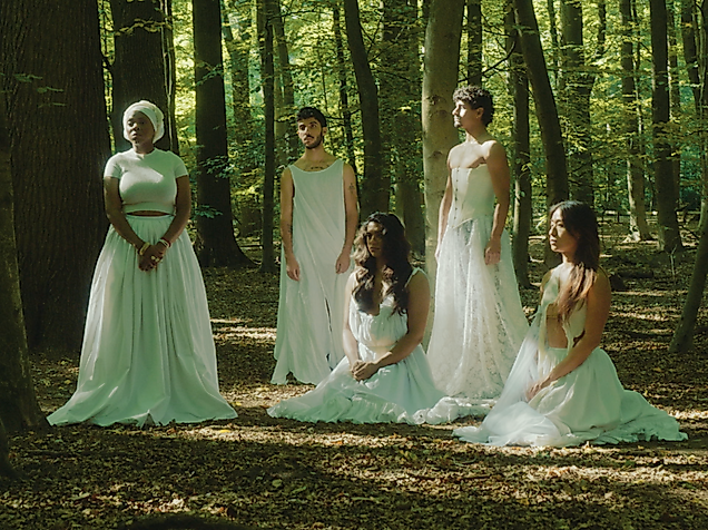 Five performers in long white dresses pose in the forest. Three of them are standing, two are sitting on the ground. None of them are looking at the camera.