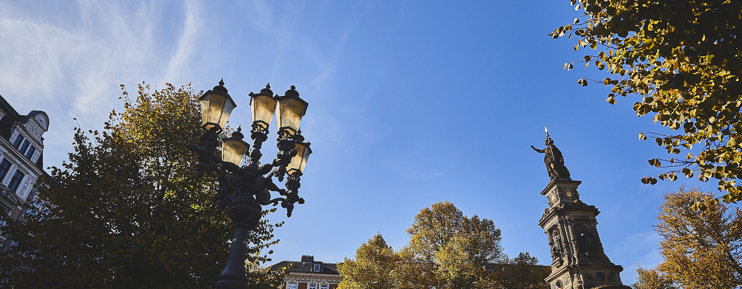 Laterne und St.-Georg-Brunnen auf dem Hansaplatz in Hamburg bei blauem Himmel und herbstlich belaubten Bäumen