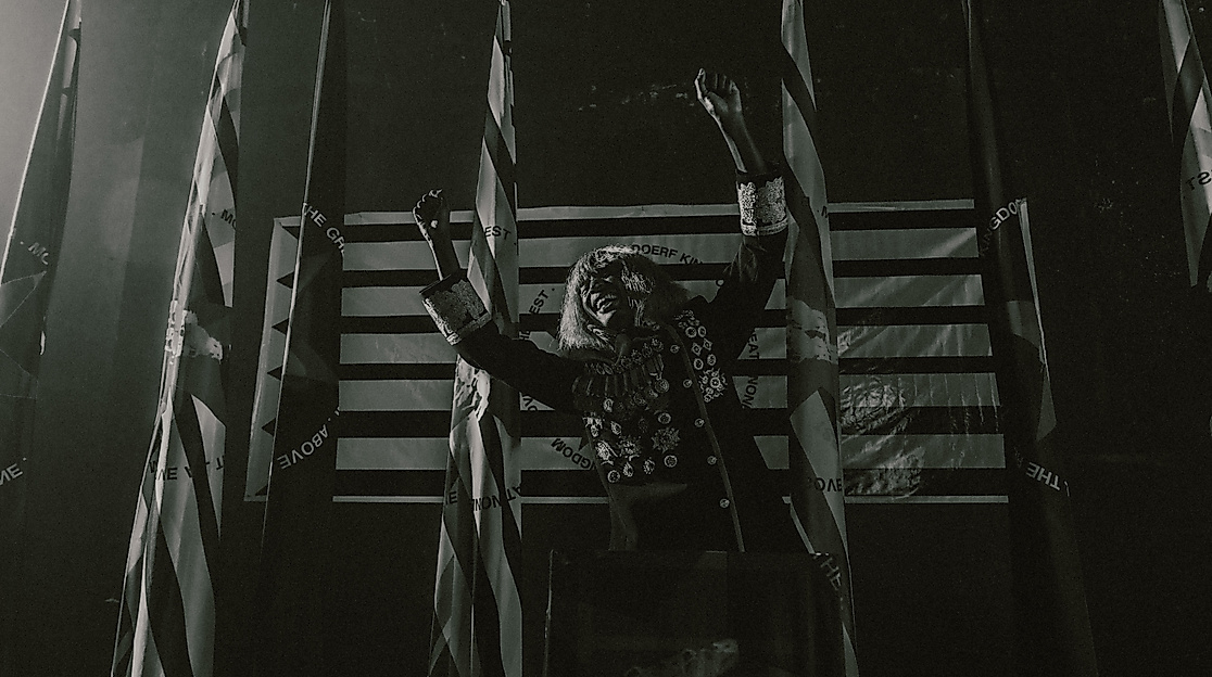 KABEAUSHÉ stands on a stage in front of four flags, wearing a uniform decorated with many medals, stretching his arms sideways and laughing.