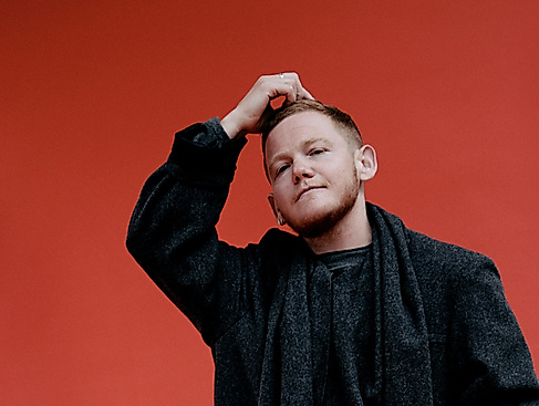 Kae Tempest stands dressed in black in front of a colorful wall with one hand on their head.