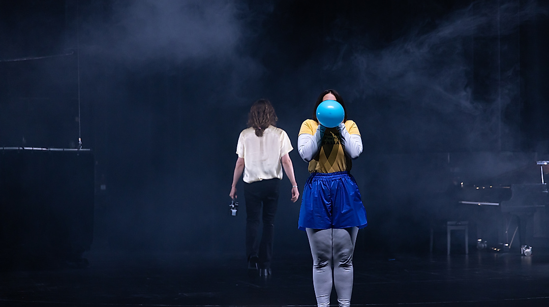 Excerpt from “My Kingdom for a Joke”: A person wearing silver leggings, blue sweatpants, and a yellow shirt holds a blue balloon in front of their face. In the background, a person dressed in black and white walks away.