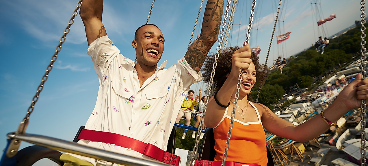 Zwei junge Menschen fahren das Kettenkarussell auf dem Hamburger DOM und halten euphorisch die Arme nach oben.