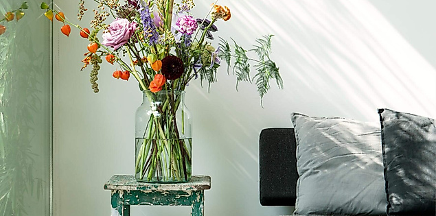Großes Blumenarrangement in Glasvase auf Hocker, daneben moderne Sitzbank im sonnendurchfluteten Raum