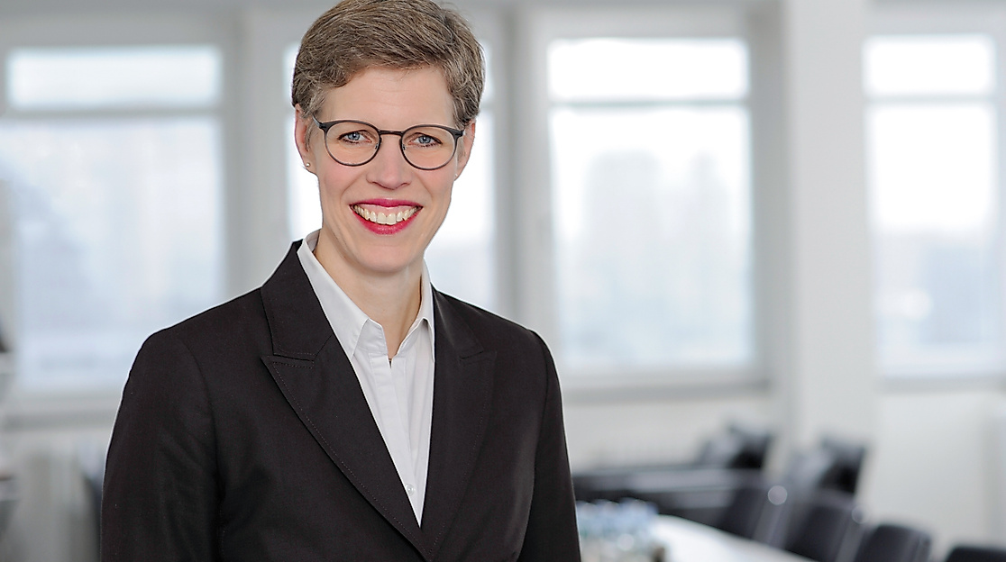 Porträt einer lächelnden Frau mit Kurzhaarfrisur und Brille im dunklen Blazer, vor hellem Konferenzraum – Ansprechpartnerin der Hamburg Tourismus GmbH
