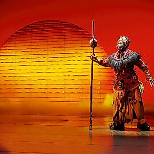 Szenenbild König der Löwen: Charakter Rafiki in afrikanischem Kostüm und Speer  bei Sonnenuntergang