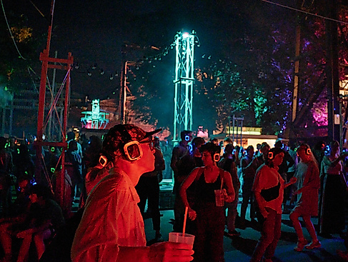 Nächtliche JAJAJA Veranstaltung mit einer Menschenmenge, die leuchtende Silent-Disco-Kopfhörer in Rot und Blau trägt. Im Vordergrund eine Person mit Cap und eckiger Brille, ein Getränk in der Hand. Im Hintergrund ist ein beleuchteter Stahlturm.