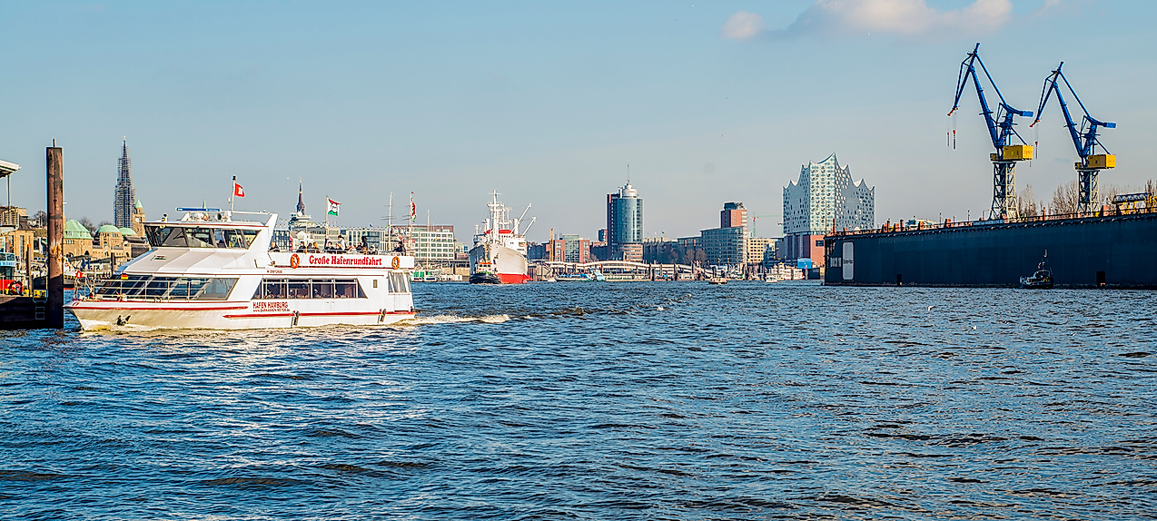 Barkasse auf der Elbe mit Blick auf Hafenkräne und Skyline von Hamburg bei Sonnenschein.
