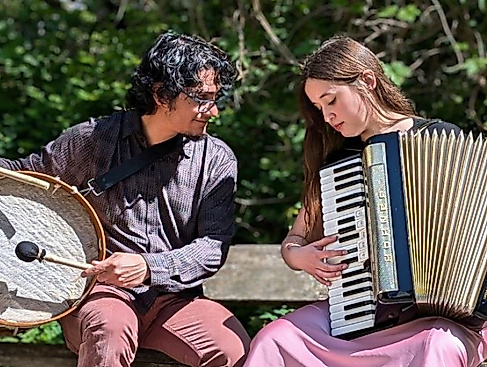 Víctor Gutiérrez und Isidora Caroca sitzen auf einer Bank. Während Víctor ein Schlaginstrument hält, hat Isidora ein Akkordeon.