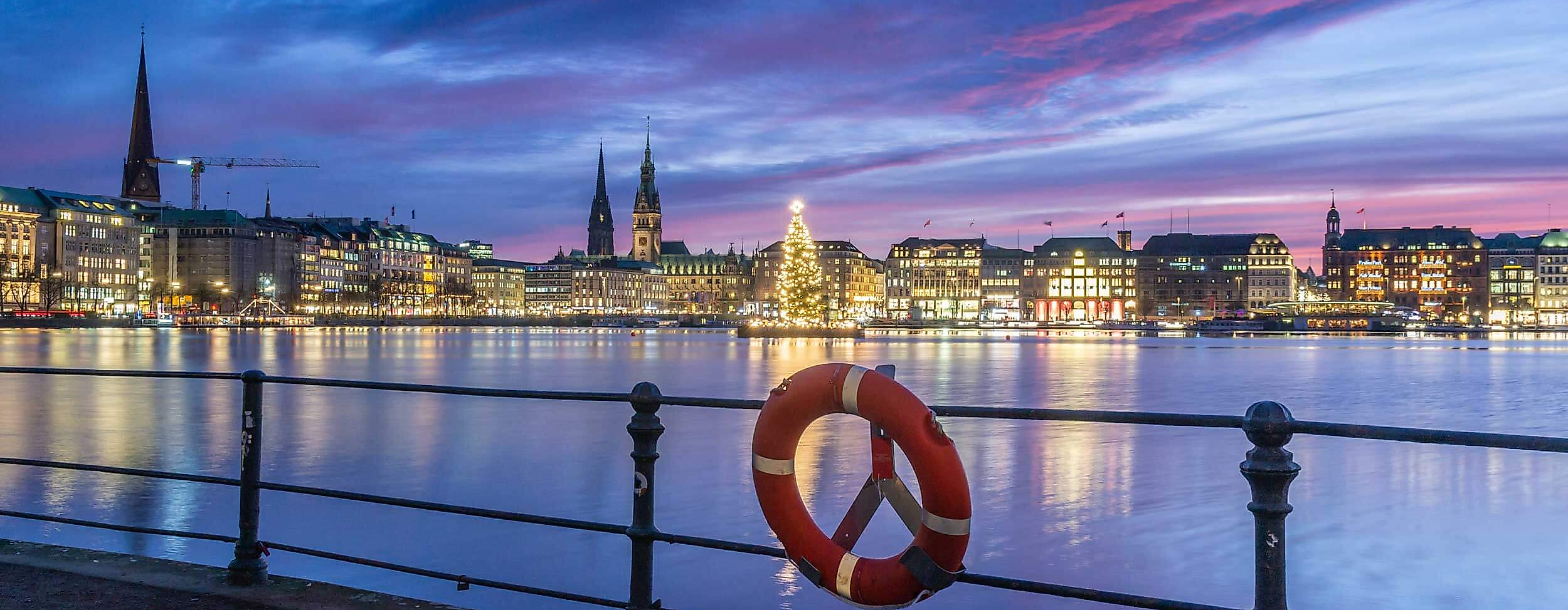 Weihnachtsbaum auf der Alster vor beleuchtetem Jungfernstieg bei blau-violettem Abendhimmel und Rettungsring im Vordergrund