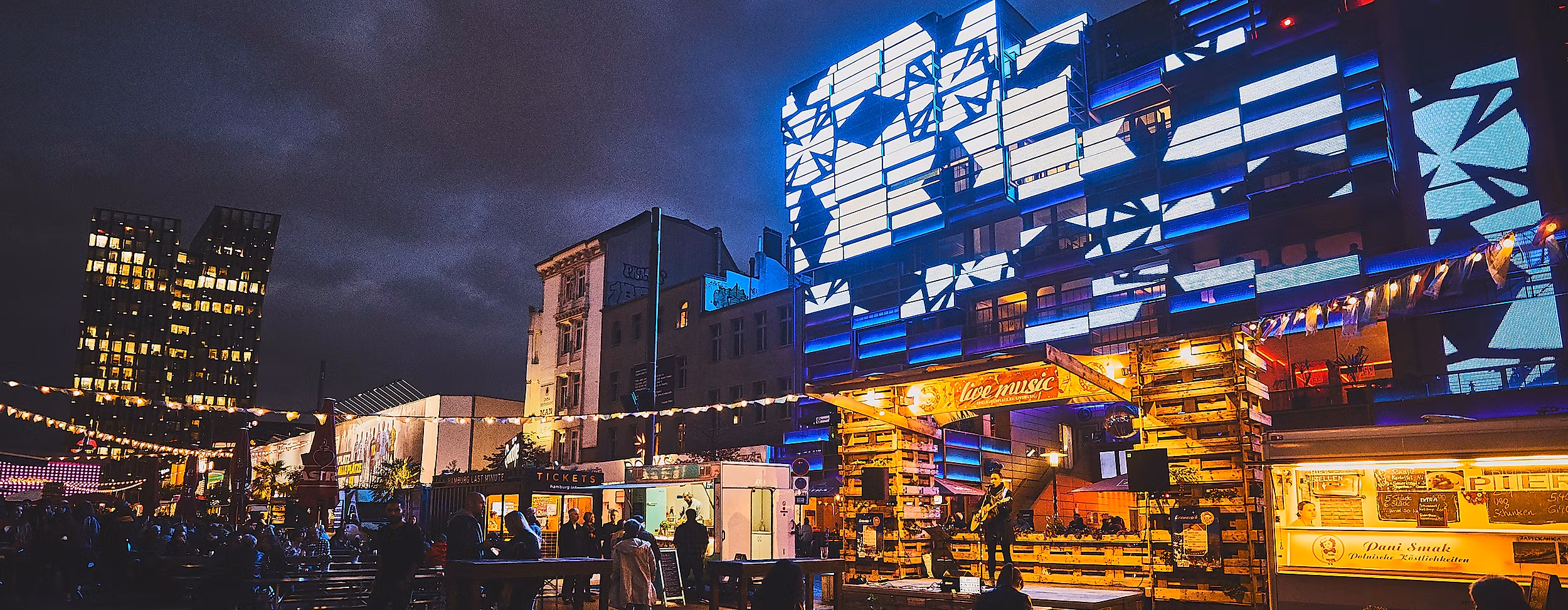 Spielbudenplatz bei Nacht mit beleuchteten Fassaden, Food-Ständen und leuchtender Straßenkunst in Blau und Gelb