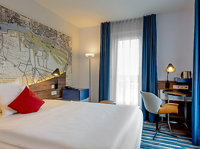 Helles Hotelzimmer mit weißem Bett, Schreibtisch, blauen Vorhängen.