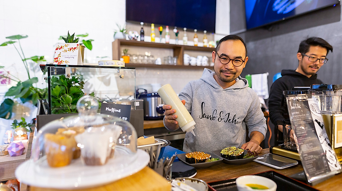 Zwei Köche bereiten asiatische Speisen an einer modernen Theke zu, im Vordergrund Kuchen unter Glasglocke