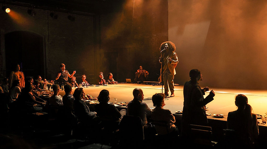 A moody theatre scene: people sit at a long table filled with food while a person on stage speaks or sings into a microphone. Warm light, intimate atmosphere, sense of community.