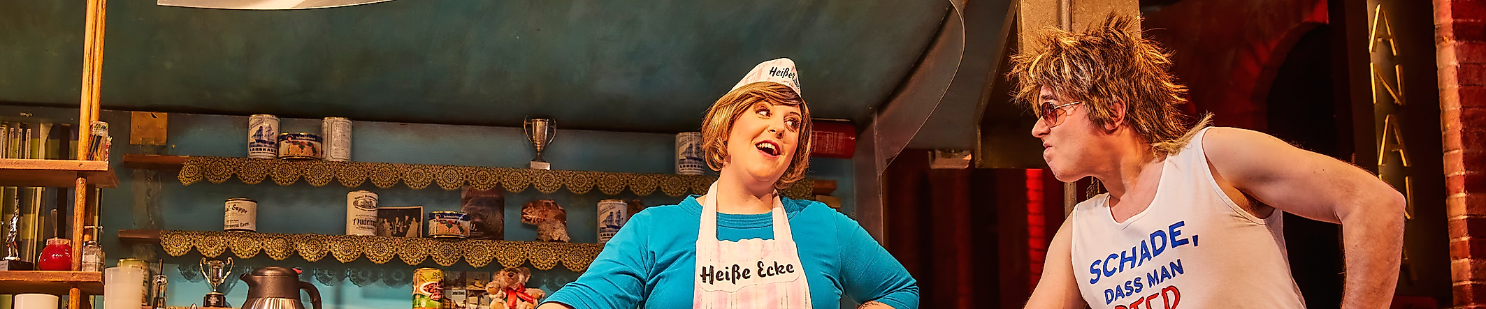 Szenenbild Heiße Ecke: Ein Mann vor dem Tresen redet mit einer Frau hinter dem Tresen