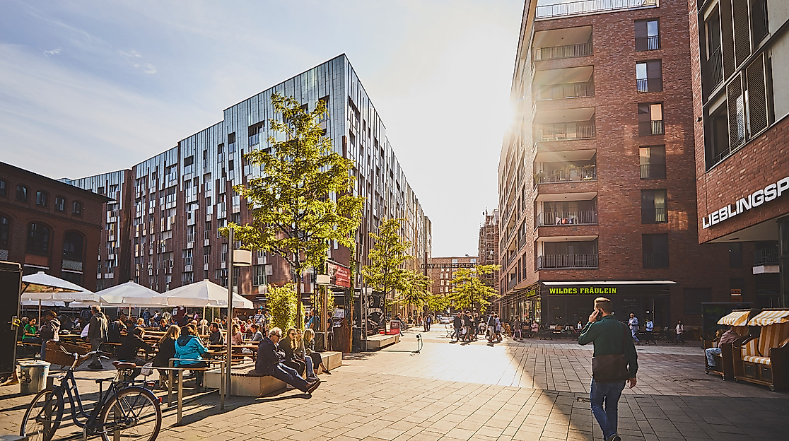 Menschen auf dem Überseeboulevard in der HafenCity Hamburg mit Cafés, Bäumen und moderner Architektur bei Sonnenschein