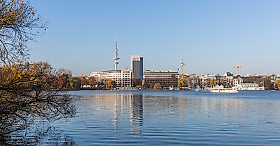 Blick über die Außenalster in Hamburg mit Uferbäumen, Segelboot und markanter Stadtsilhouette im Sonnenlicht.