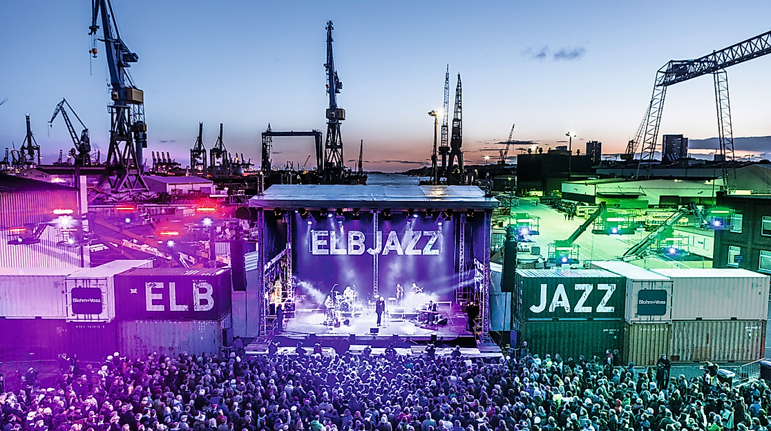 Beleuchtete Bühne des ELBJAZZ Festivals im Hamburger Hafen mit Menschenmenge und Hafenkränen bei Abenddämmerung.