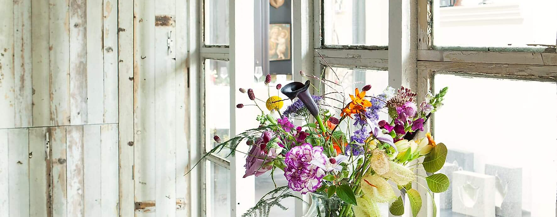 Vase mit frischen, bunt gemischten Blumen auf Holztisch vor Fensterfront in hellem Kreativraum