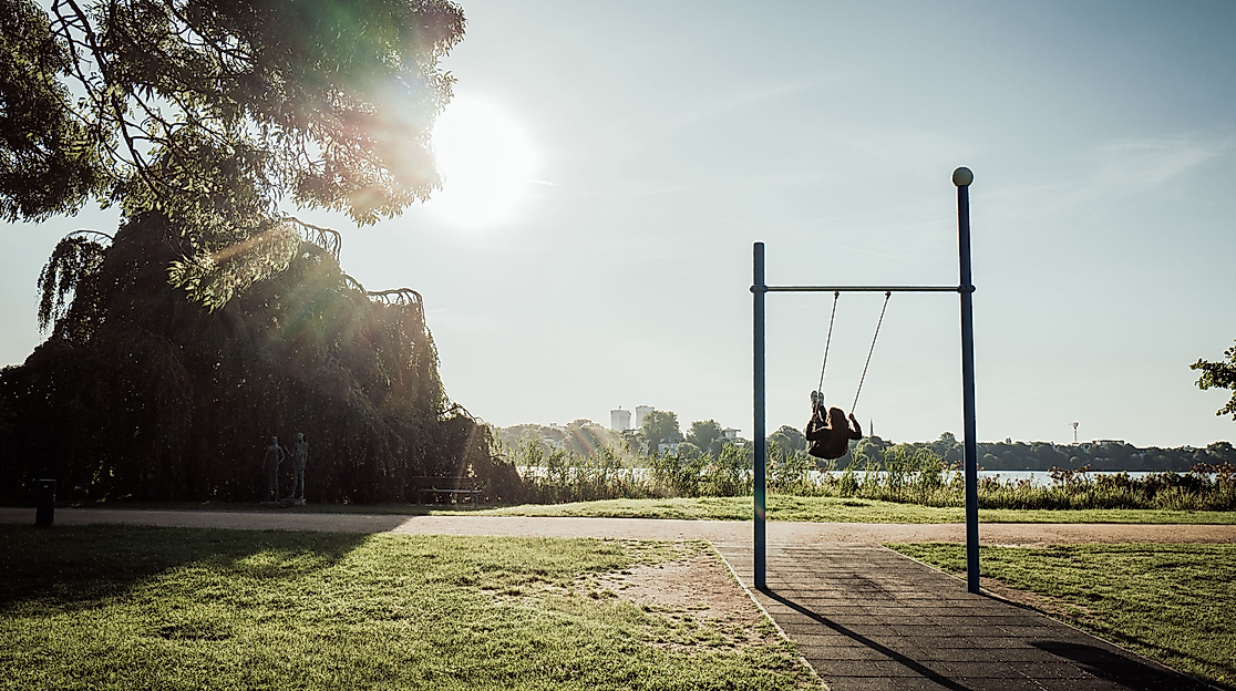 Person schaukelt im Alsterpark bei Sonnenschein, mit Blick auf die Außenalster und umliegende Natur.