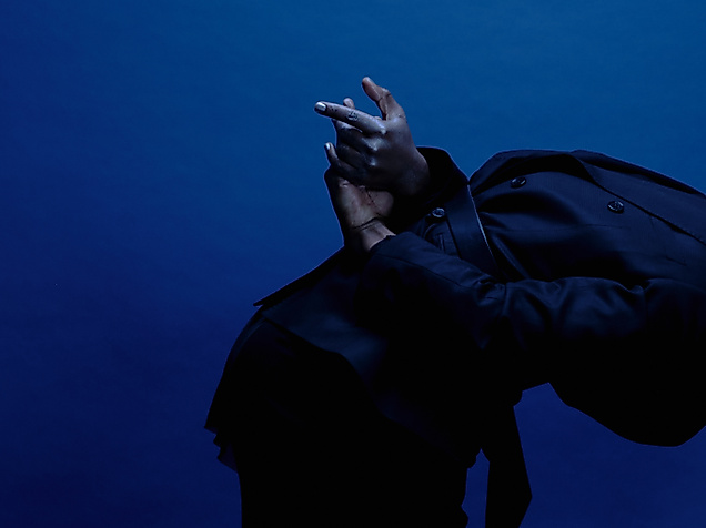 A performer is seen from the side, leaning forward, a black hoodie pulled over their head. Their hands are behind their back and stretched out.