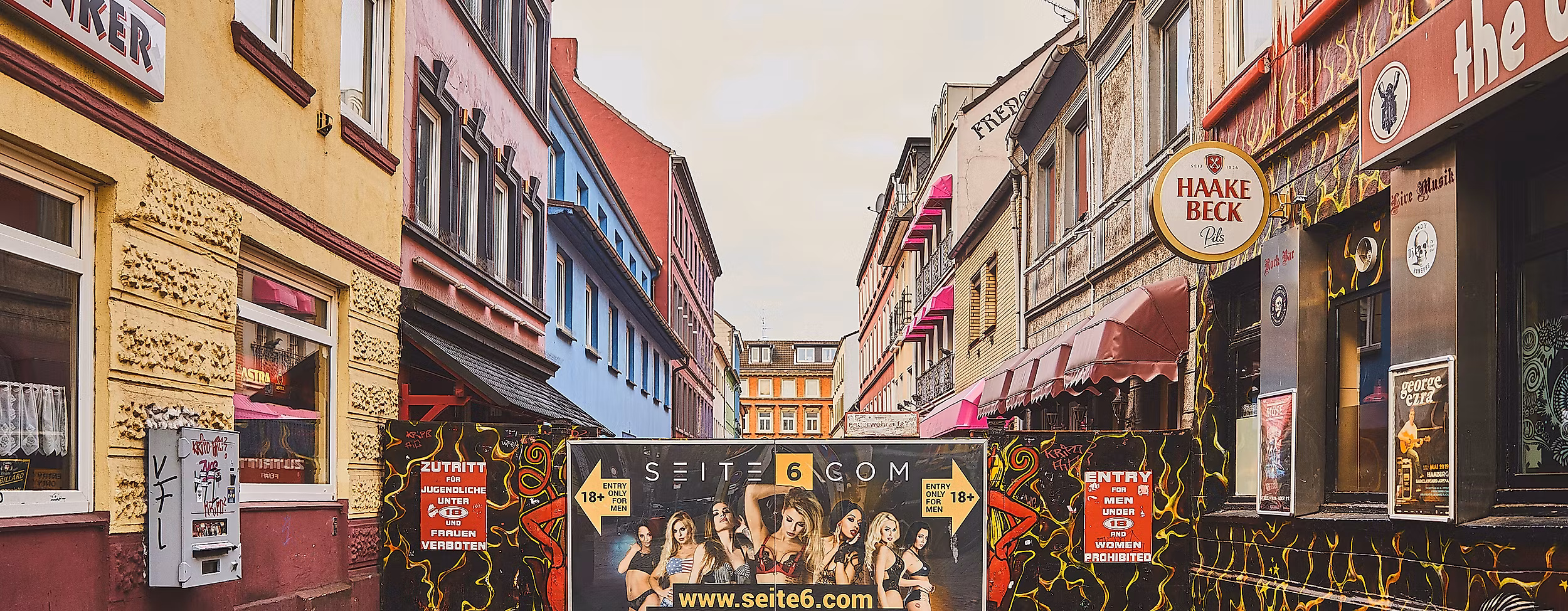 Blick in die Herbertstraße mit Sichtschutzwand, flankiert von bunt beleuchteten Fassaden und Schildern auf der Reeperbahn