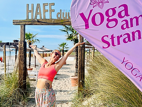 Frau steht am Strand vor den HAFFdüün bei Sonne und mit einem Schild Strandyoga YOGACOAST in Haffkrug an der Ostsee