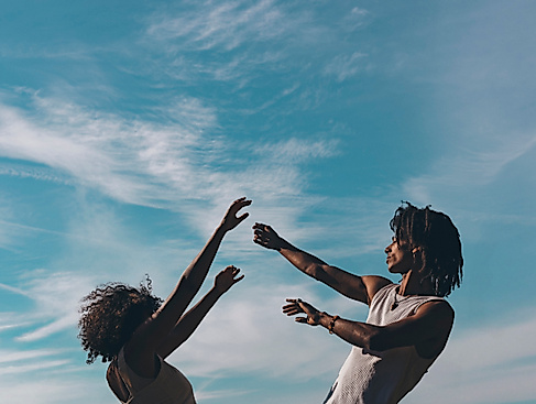 Two people raise their arms toward the sky, silhouetted against a blue sky with wispy clouds.