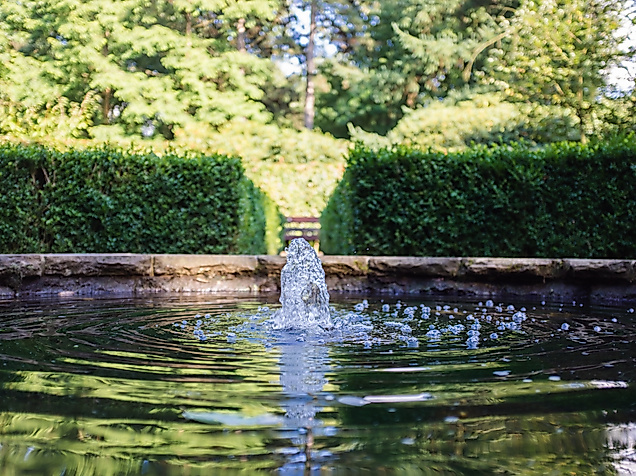 Wasser sprudelt aus einem Brunnenbecken, eingefasst von Hecken und Bäumen in einer ruhigen Parkanlage.