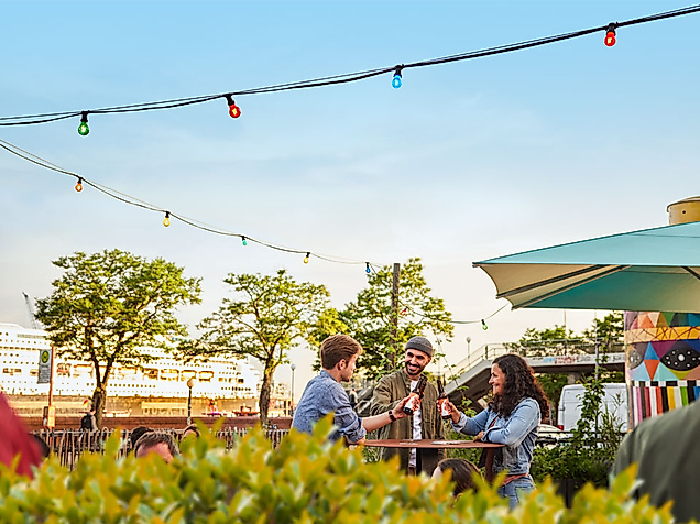 Menschen trinken Bier an Stehtischen unter Lichterketten bei Sonnenuntergang auf der Hamburg Beer Week