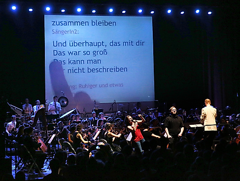 Ein Orchester spielt auf einer Bühne, im Hintergrund ist eine große Leinwand, auf die ein Songtext projiziert wird. Vor dem Orchester steht eine junge Frau, die fröhlich in ein Mikrofon singt.
