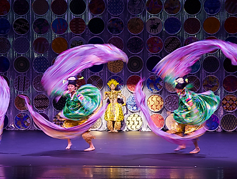Two dancers in colourful costumes whirl bright ribbons, with one person in gold costume standing behind them in front of circular ornaments.