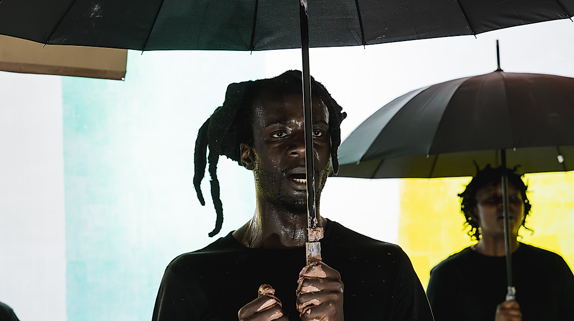 Excerpt from the production DAMBUDZO by nora chipaumire. A person stands holding an umbrella in front of a colorful wall. Behind them, two other people are visible in the background.