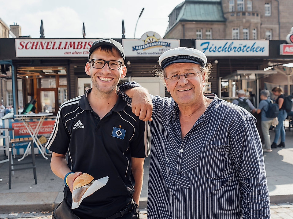 Zwei Männer vor Fischbrötchenständen an den Landungsbrücken, einer trägt Rikscha-Fahrer-Shirt